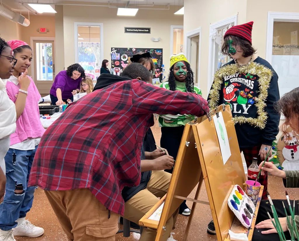 People browsing artwork in a bright classroom, with one person leaning over an easel display.