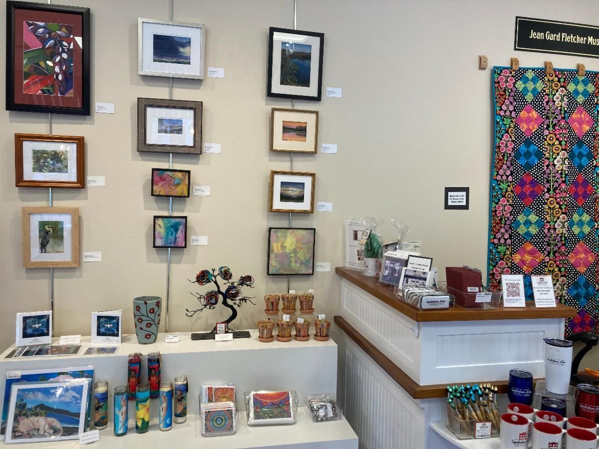 Art gallery display with colorful framed artwork, cards, crafts, and a wooden counter