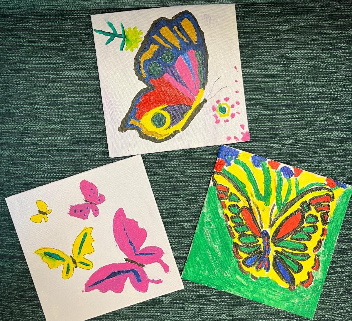 Three colorful butterfly drawings on small square papers arranged on a textured green surface