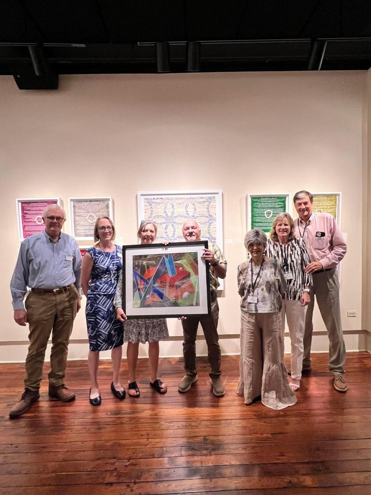 Five people posing with framed artwork in a gallery, with polished wood floor and wall-mounted pieces behind them.