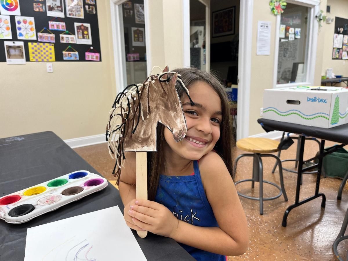 Child smiling and holding a paintbrush and paper craft in an art classroom, with watercolor tray nearby