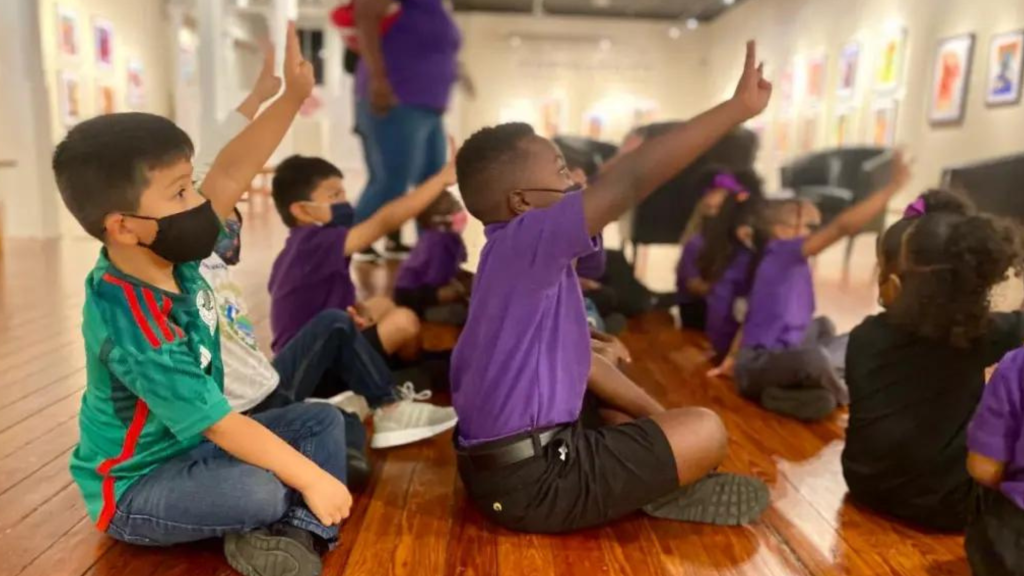 Children in purple and green shirts sit on a wooden floor, raising their hands in an art gallery.
