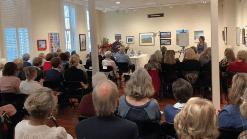 Audience seated in an art gallery listening to a speaker at the front of the room
