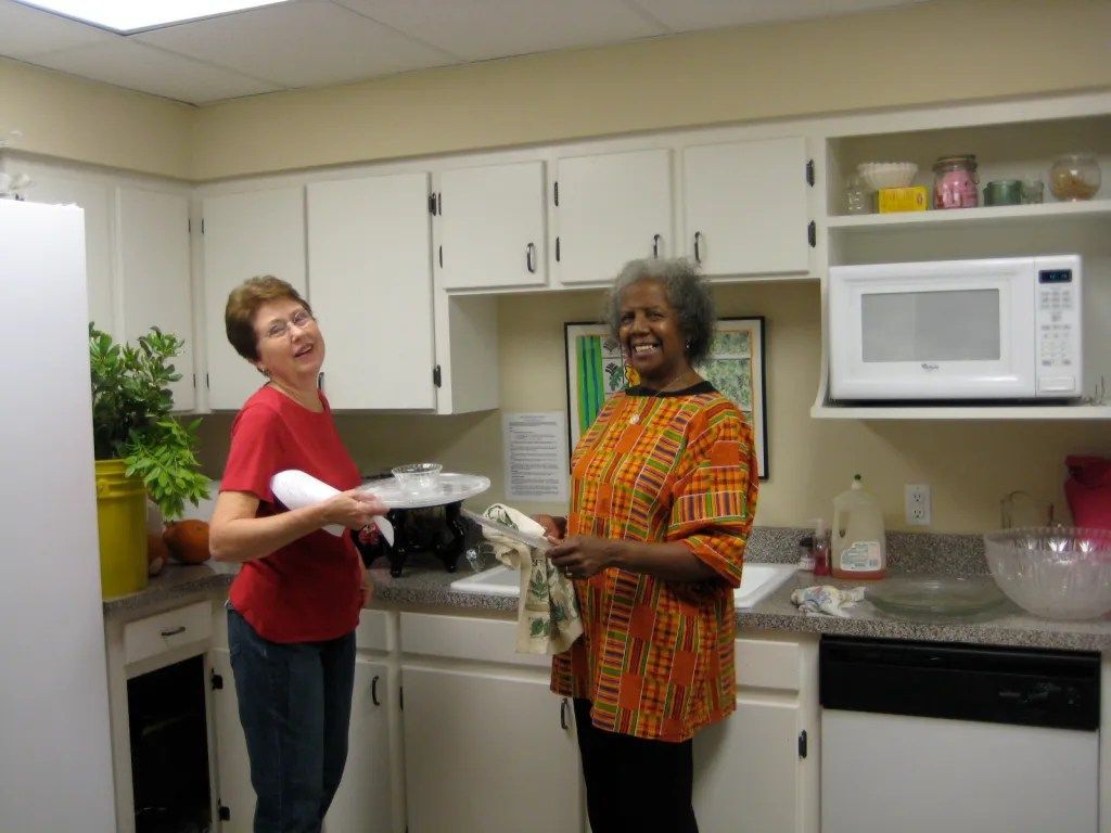 Two women smiling in a kitchen, one holding a plate and the other holding a dish towel.
