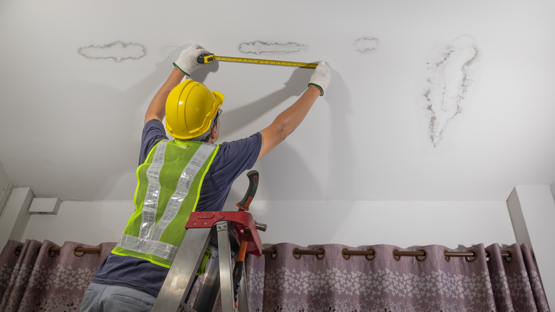 Worker in yellow hard hat painting a white ceiling above curtains using a roller on a ladder