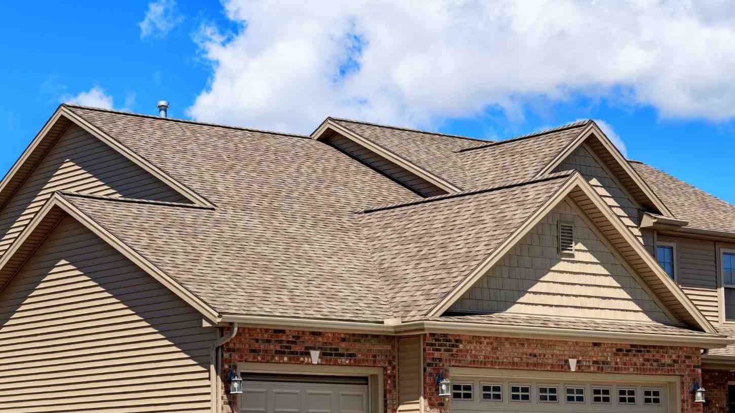 Suburban house roof with tan shingles and multiple gables under a blue sky