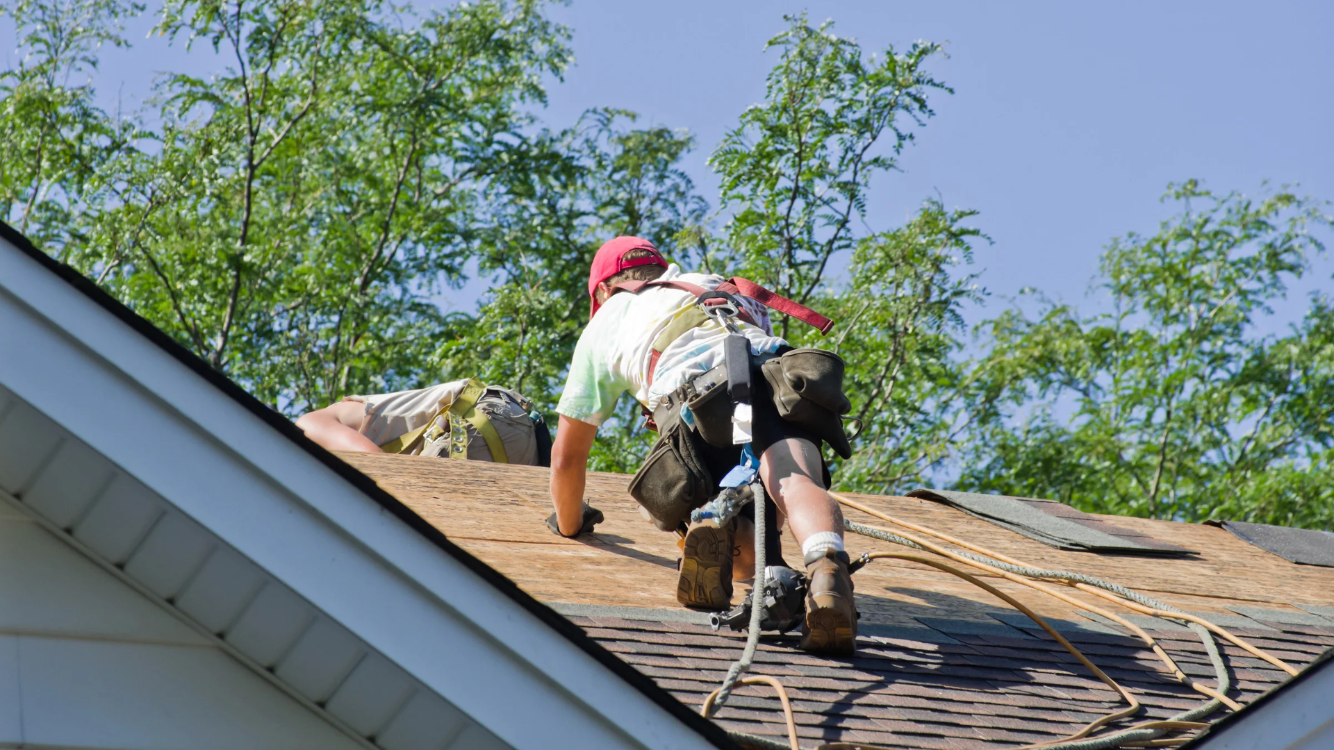 Worker on a steep roof installing shingles beside a white gutter under a blue sky