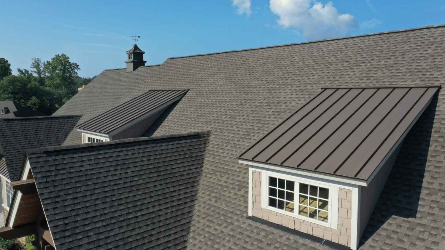 Aerial view of a gray shingle roof with two dormers and a metal-roofed windowed dormer under a blue sky