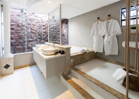 Casa de banho moderna e luminosa, com duche revestido a pedra, lavatório suspenso, banheira e roupões brancos pendurados junto às escadas