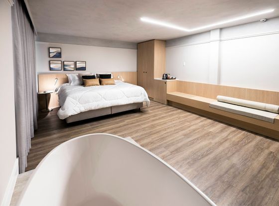Quarto moderno e minimalista, com uma cama grande, piso de madeira e um longo banco sob a janela, em meio a paredes brancas e claras.