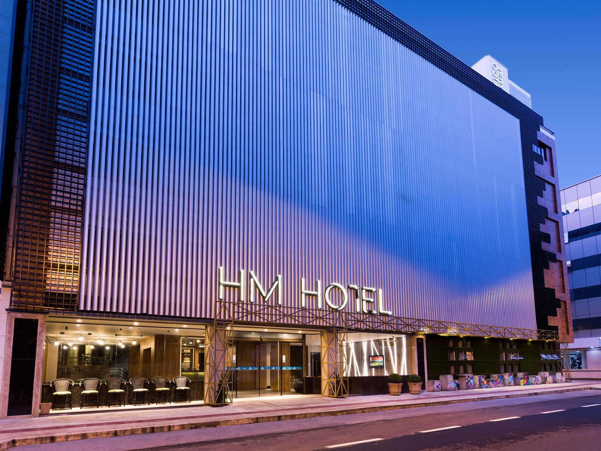 Exterior do HM Hotel, com entrada iluminada e fachada azul ondulante ao anoitecer