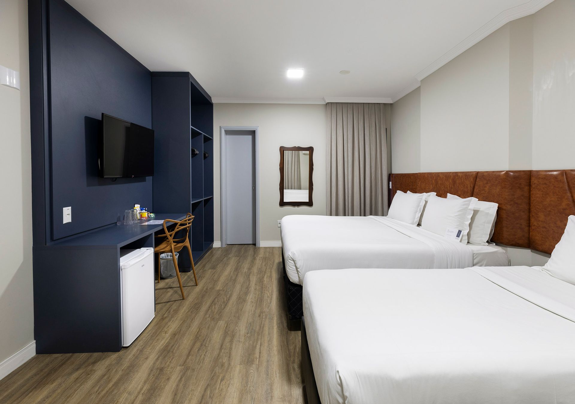 Quarto de hotel moderno com duas camas brancas, parede de destaque azul-escura, piso de madeira e TV montada na parede