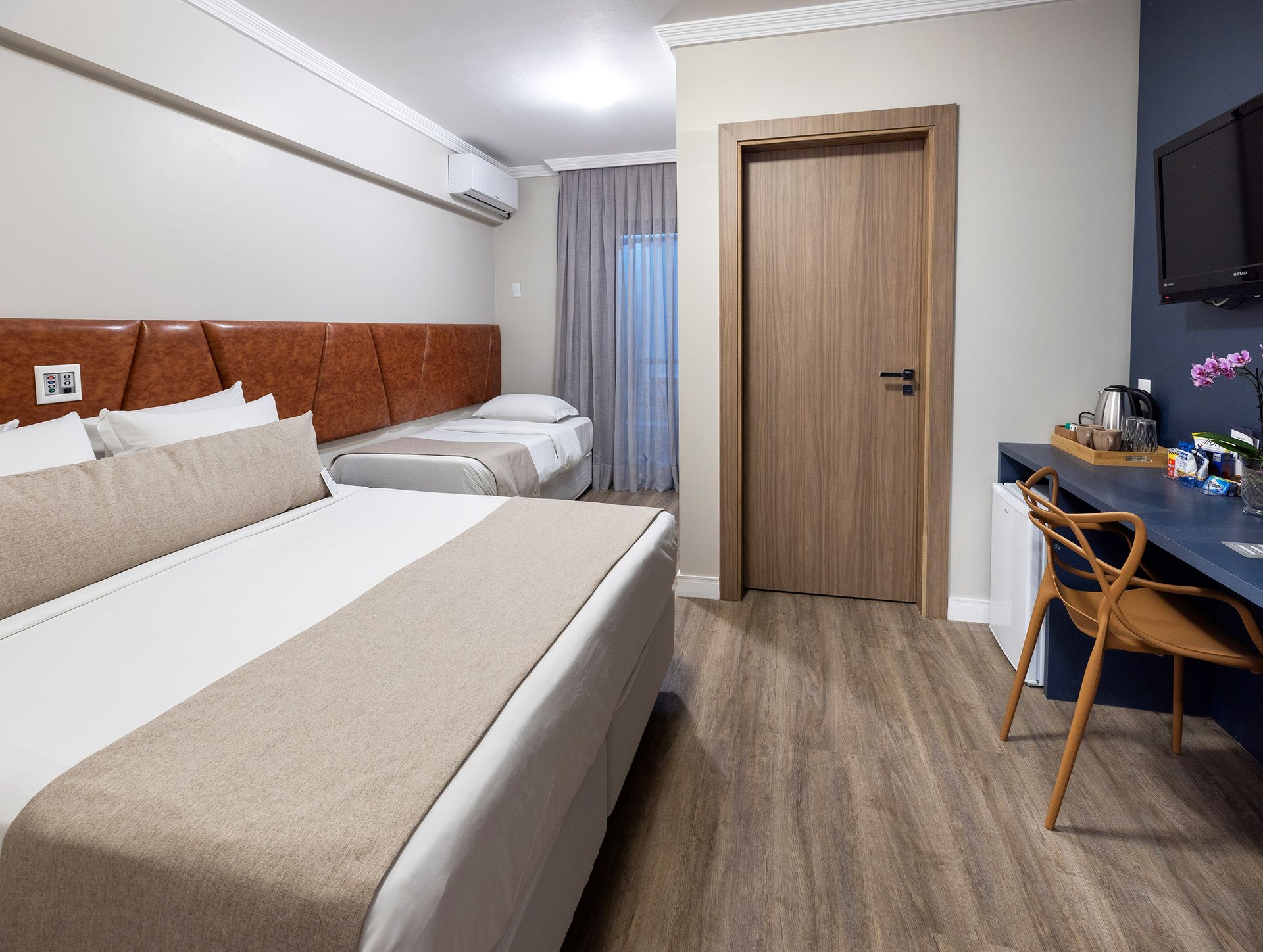 Quarto de hotel com duas camas, porta de madeira, escrivaninha, TV fixada na parede e decoração em tons neutros.