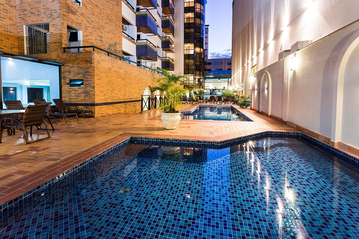 Piscina na cobertura ao lado de edifícios de tijolo e brancos ao anoitecer, com espreguiçadeiras e as luzes da cidade refletidas na água