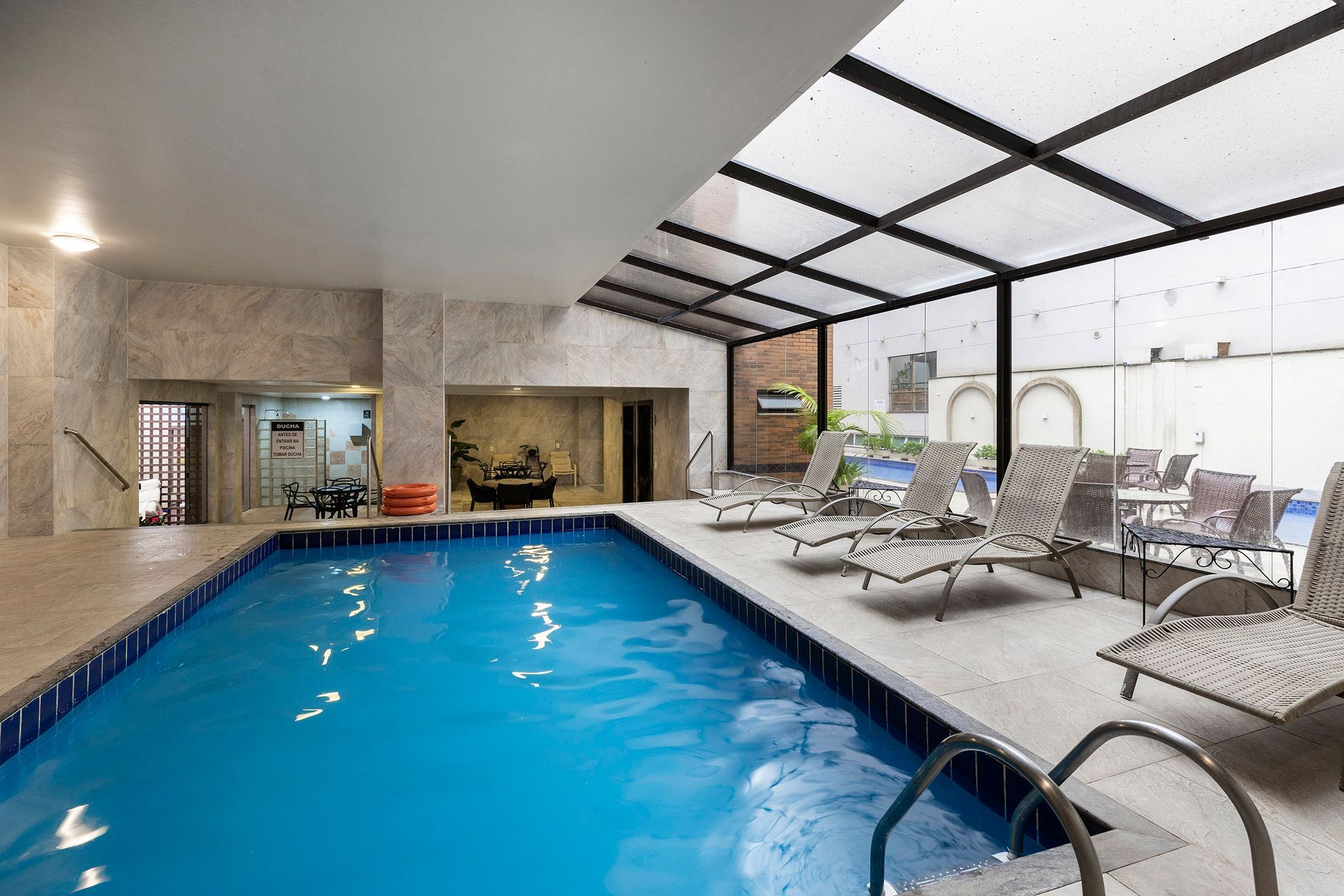 Piscina coberta com água azul, espreguiçadeiras e grandes janelas junto a um pátio coberto