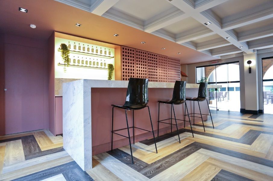 Área de bar moderna, com banquetas altas, balcão de mármore e piso de madeira com padrão geométrico
