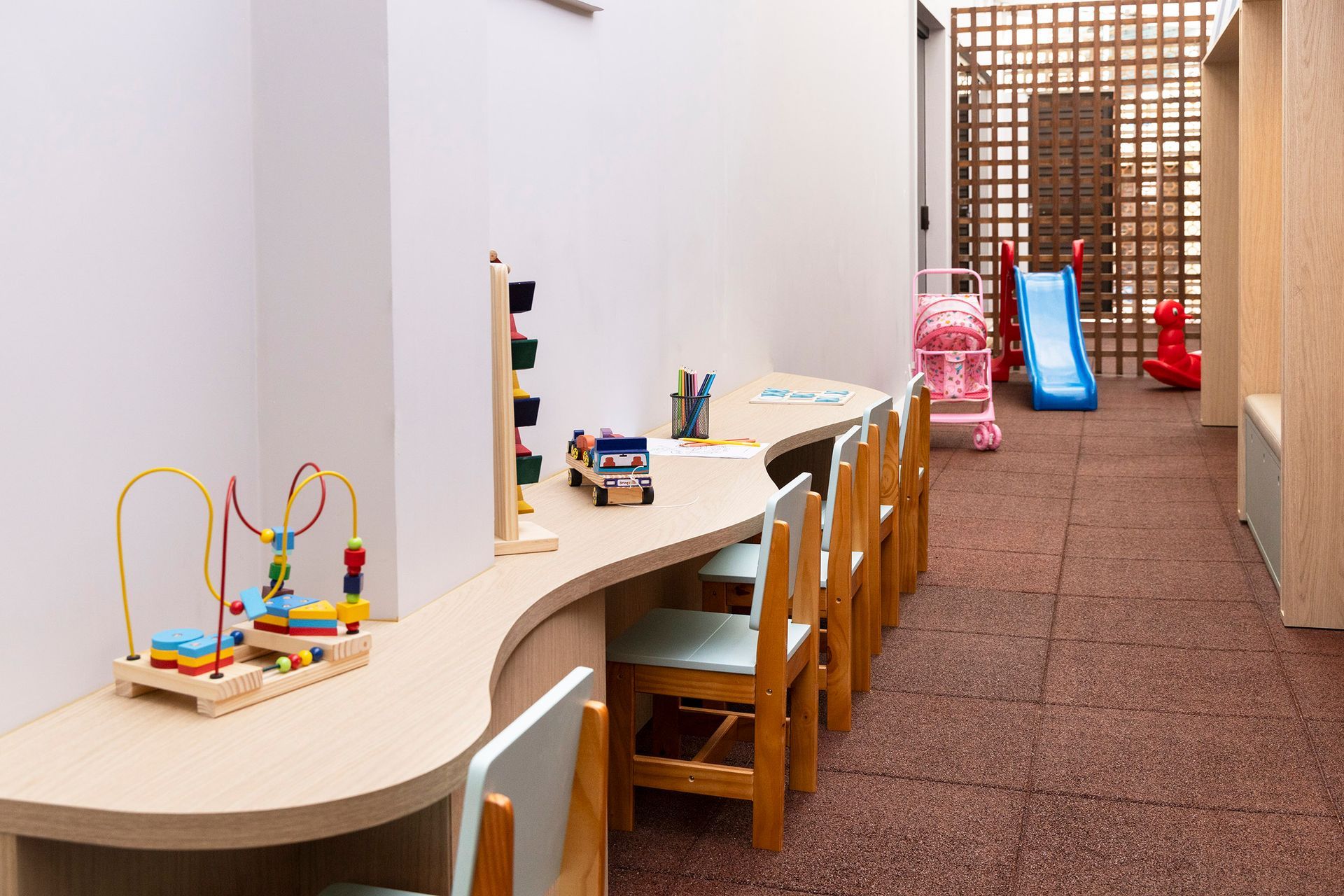 Área infantil com mesas de brinquedo, cadeiras de madeira, um escorregador e equipamentos coloridos para brincar.
