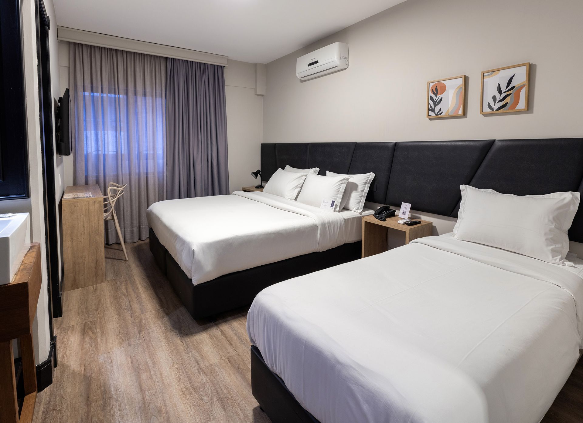 Quarto de hotel moderno com duas camas bem arrumadas, cortinas cinzentas, quadros na parede e uma pequena mesa de cabeceira