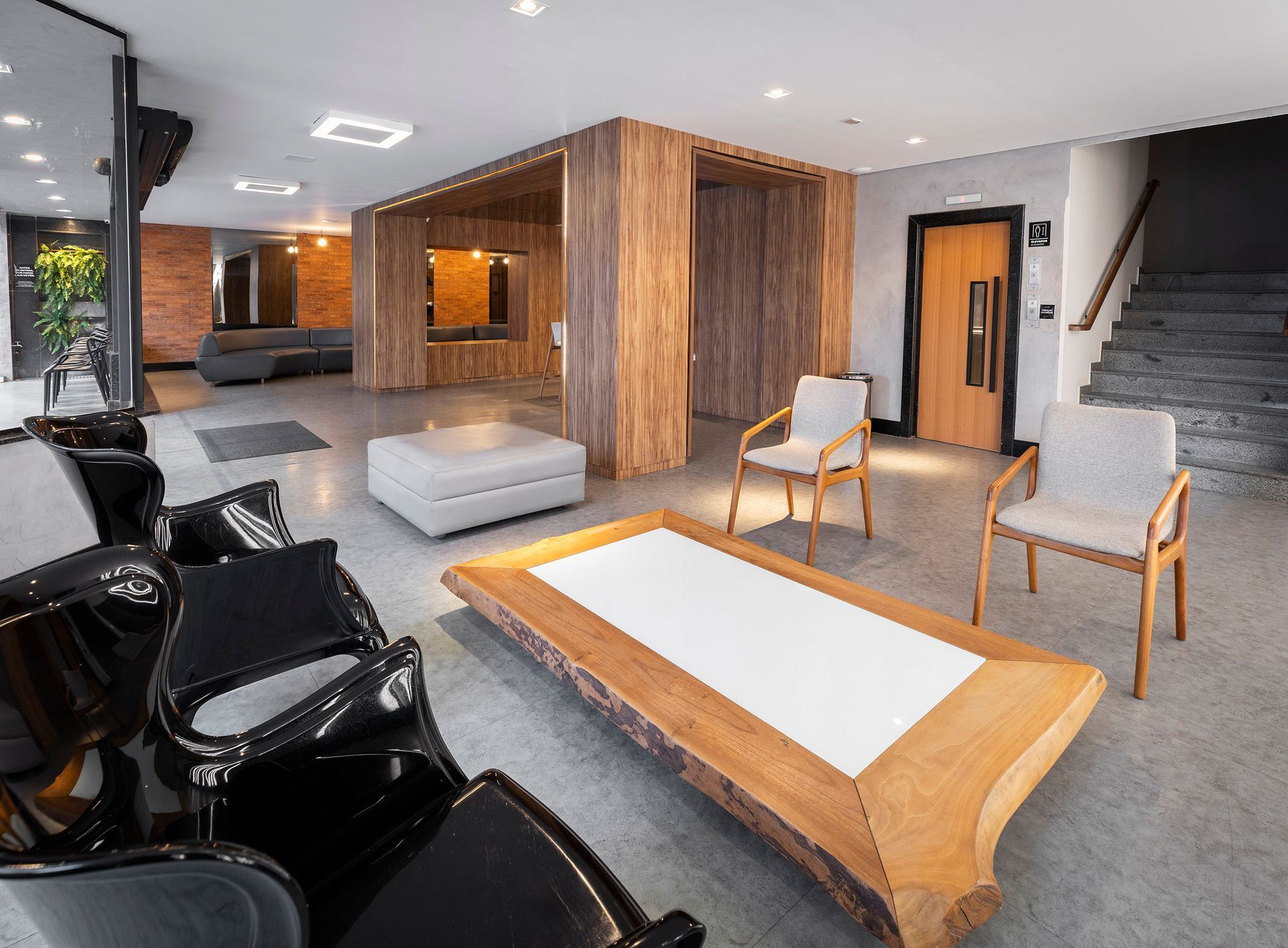 Lobby moderno com detalhes em madeira, cadeiras pretas, sofá cinza, mesa branca e escada sob iluminação intensa