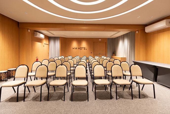 Sala de conferências vazia, com filas de cadeiras bege, paredes revestidas de madeira e um projeto de iluminação no teto