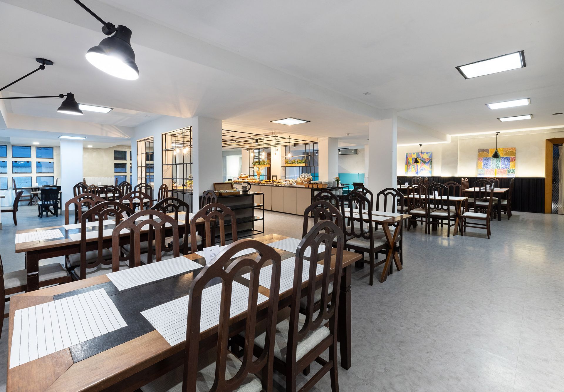Sala de jantar iluminada, com mesas e cadeiras de madeira dispostas de forma organizada e lâmpadas fluorescentes no teto