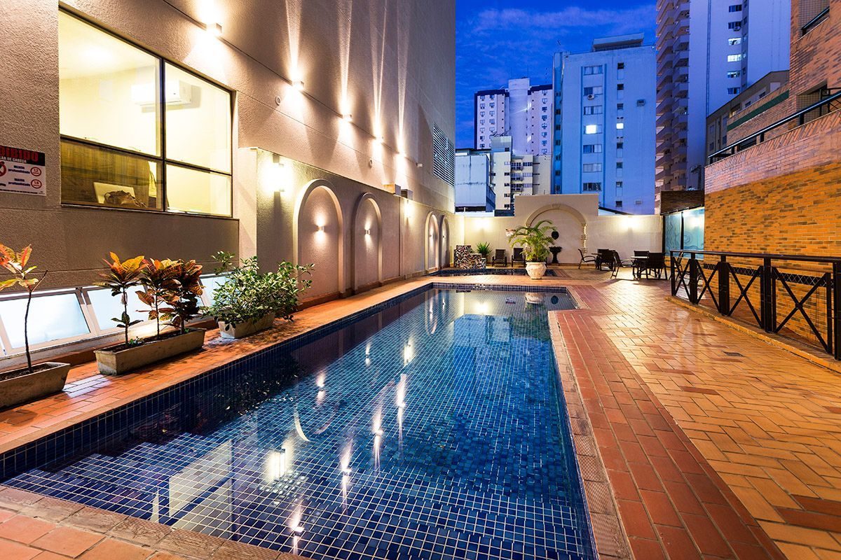 Piscina estreita na cobertura, ao lado de um edifício iluminado ao anoitecer, com torres da cidade ao fundo.