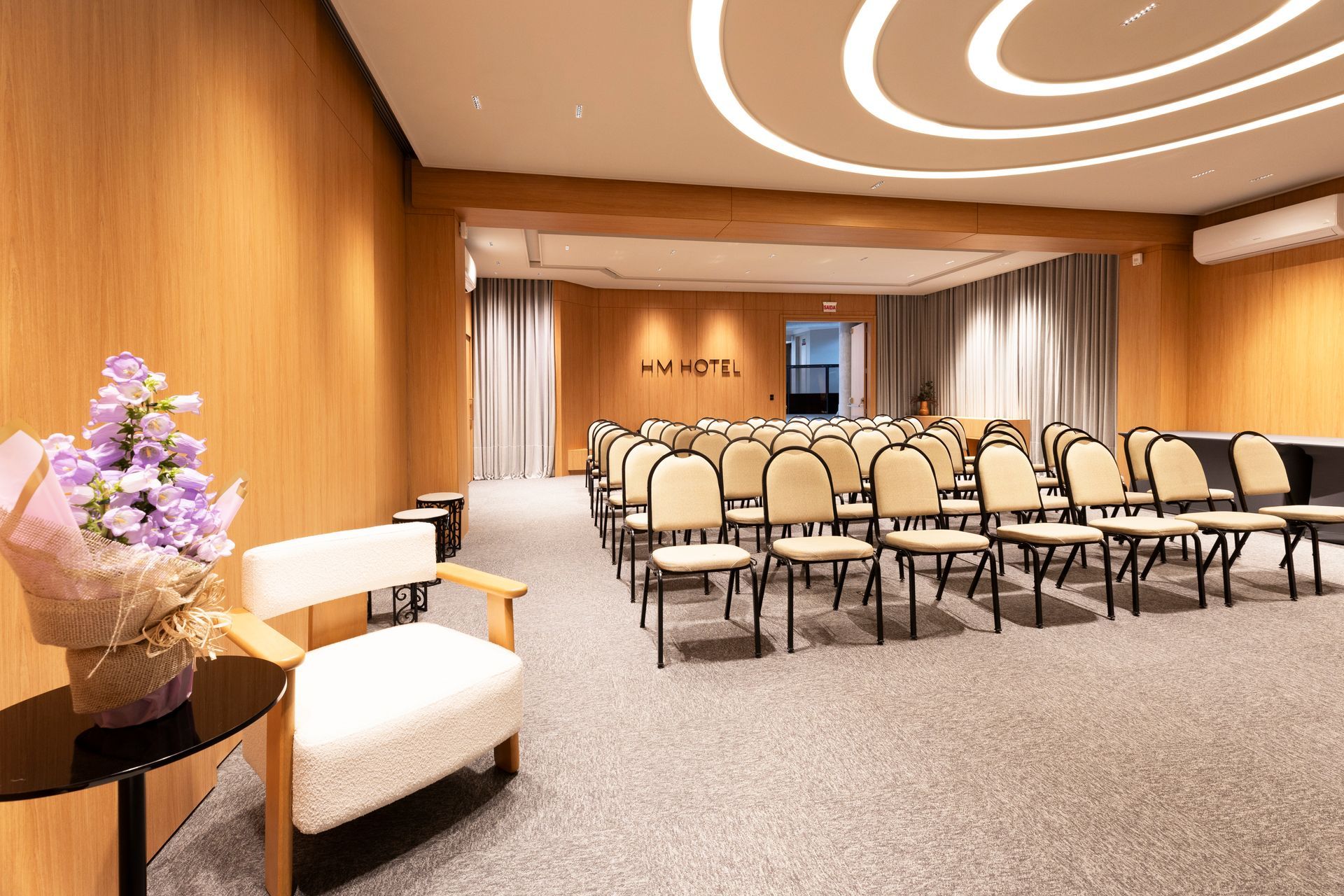 Sala de conferências com fileiras de cadeiras, paredes revestidas de madeira e luzes quentes no teto