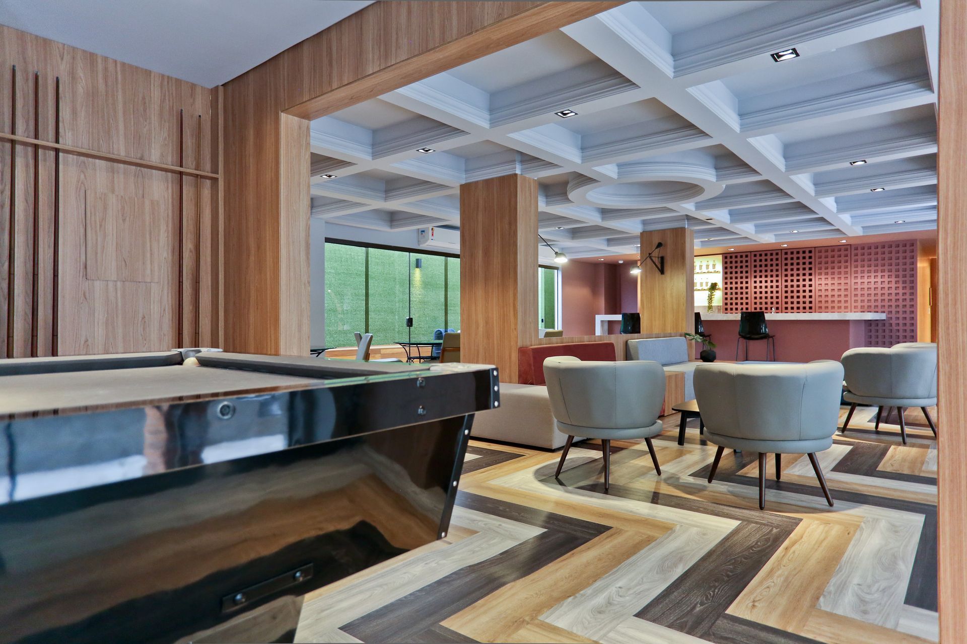 Sala de estar moderna com cadeiras azuis, tapete estampado, paredes de madeira e mesa de bilhar sob um teto em caixotões