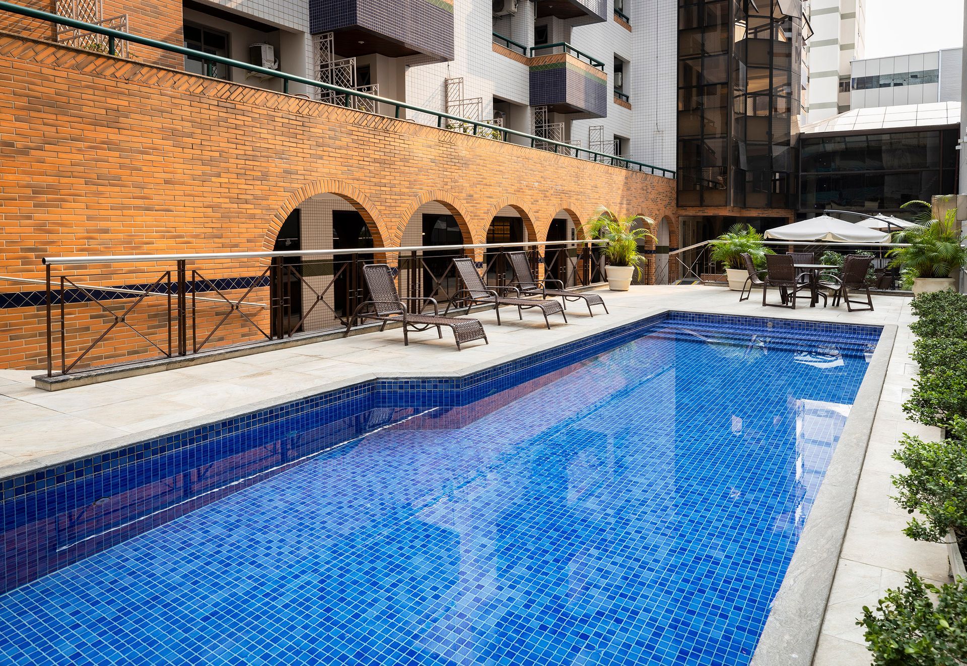 Piscina ao ar livre ao lado de um edifício de tijolos com toldos em arco e espreguiçadeiras em um dia ensolarado