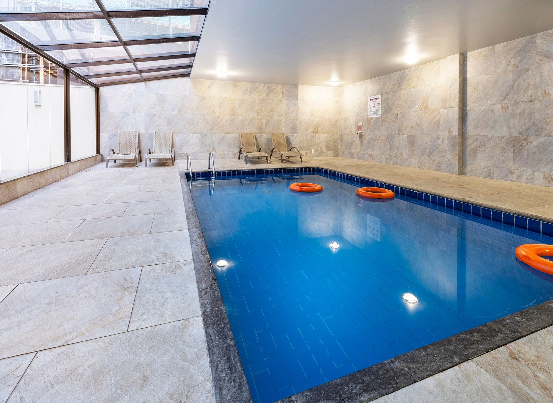 Piscina interior com água azul-viva e boias laranja ao lado de uma área de pátio coberta