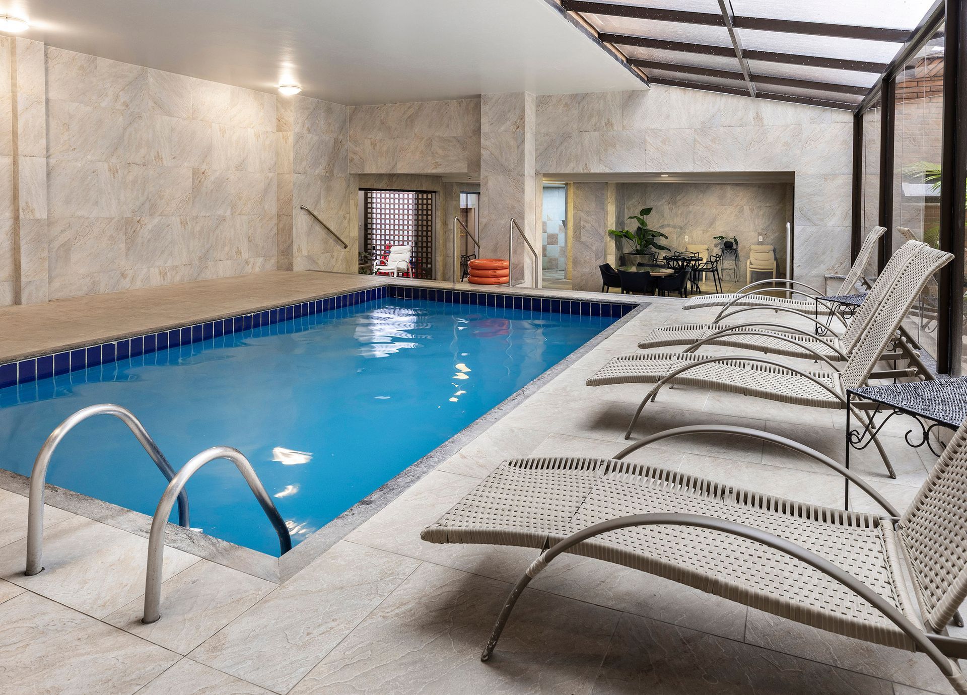 Piscina interior com água azul, escada de metal e espreguiçadeiras sob um pátio coberto