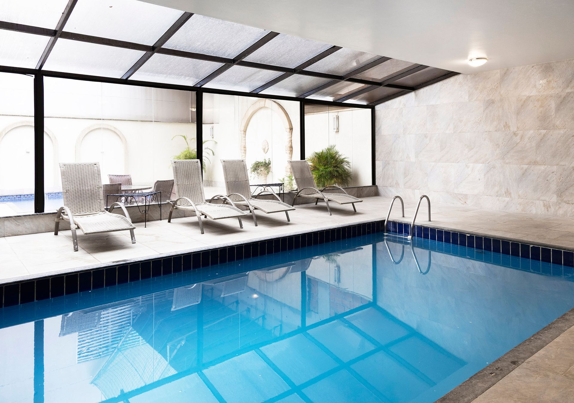 Piscina interior com água azul-viva, espreguiçadeiras e plantas ao lado de um pátio coberto