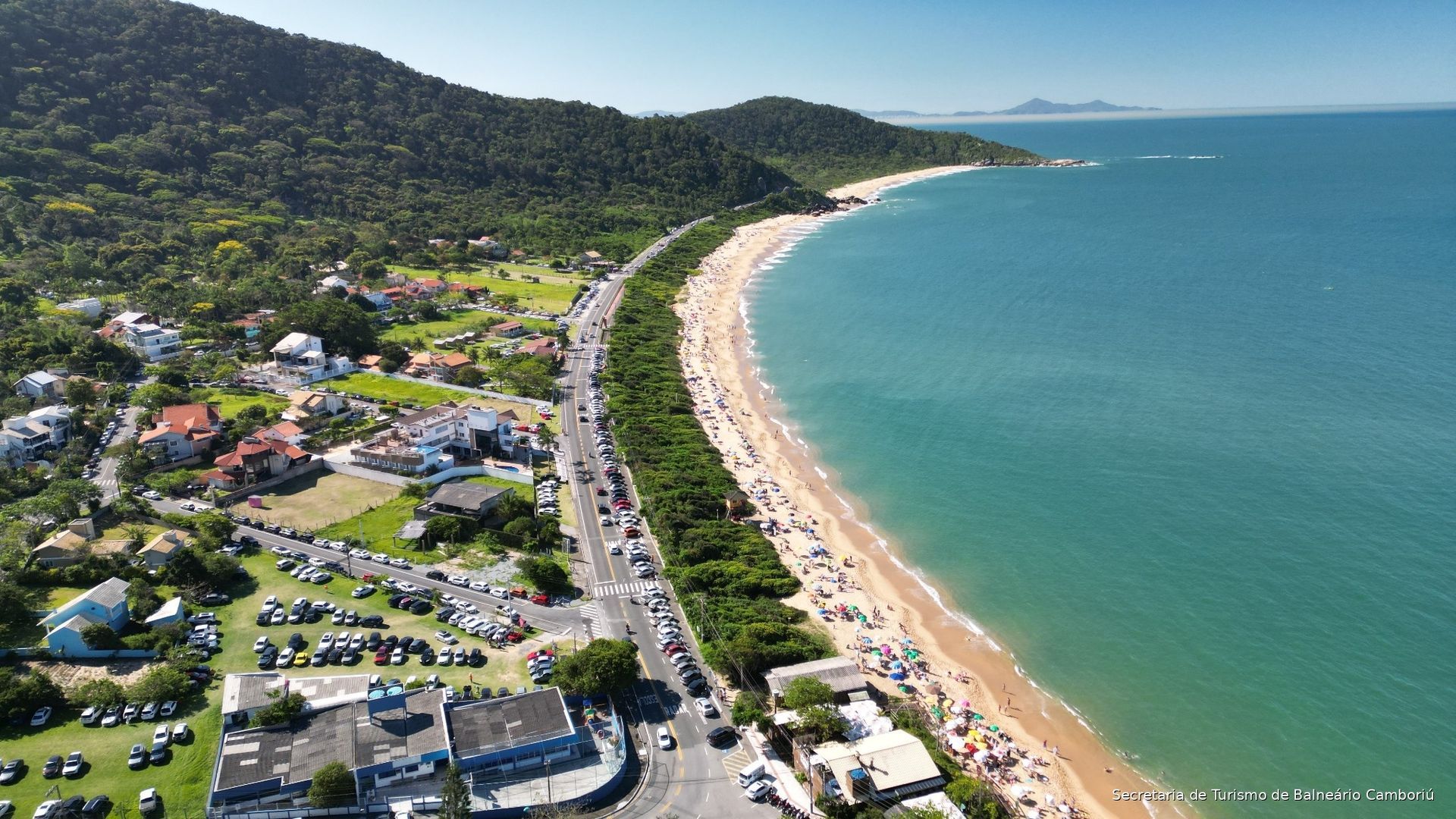 Vista aérea de uma praia de areia, mar turquesa, estrada costeira e casas ao lado de colinas verdejantes.