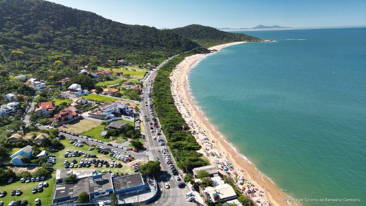 Vista aérea de uma praia de areia, mar turquesa, estrada costeira e casas ao lado de colinas verdejantes.