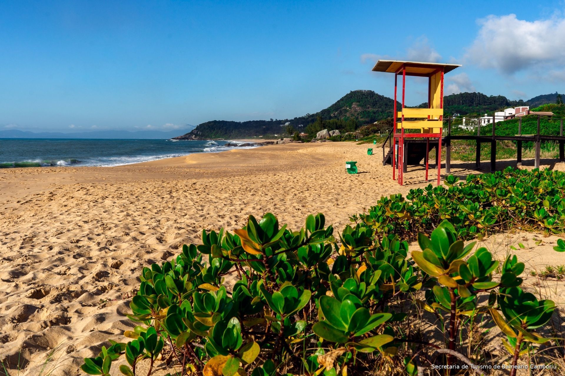 Praia ensolarada com areia dourada, oceano azul, arbustos verdes e uma torre de salva-vidas vermelha sob um céu limpo