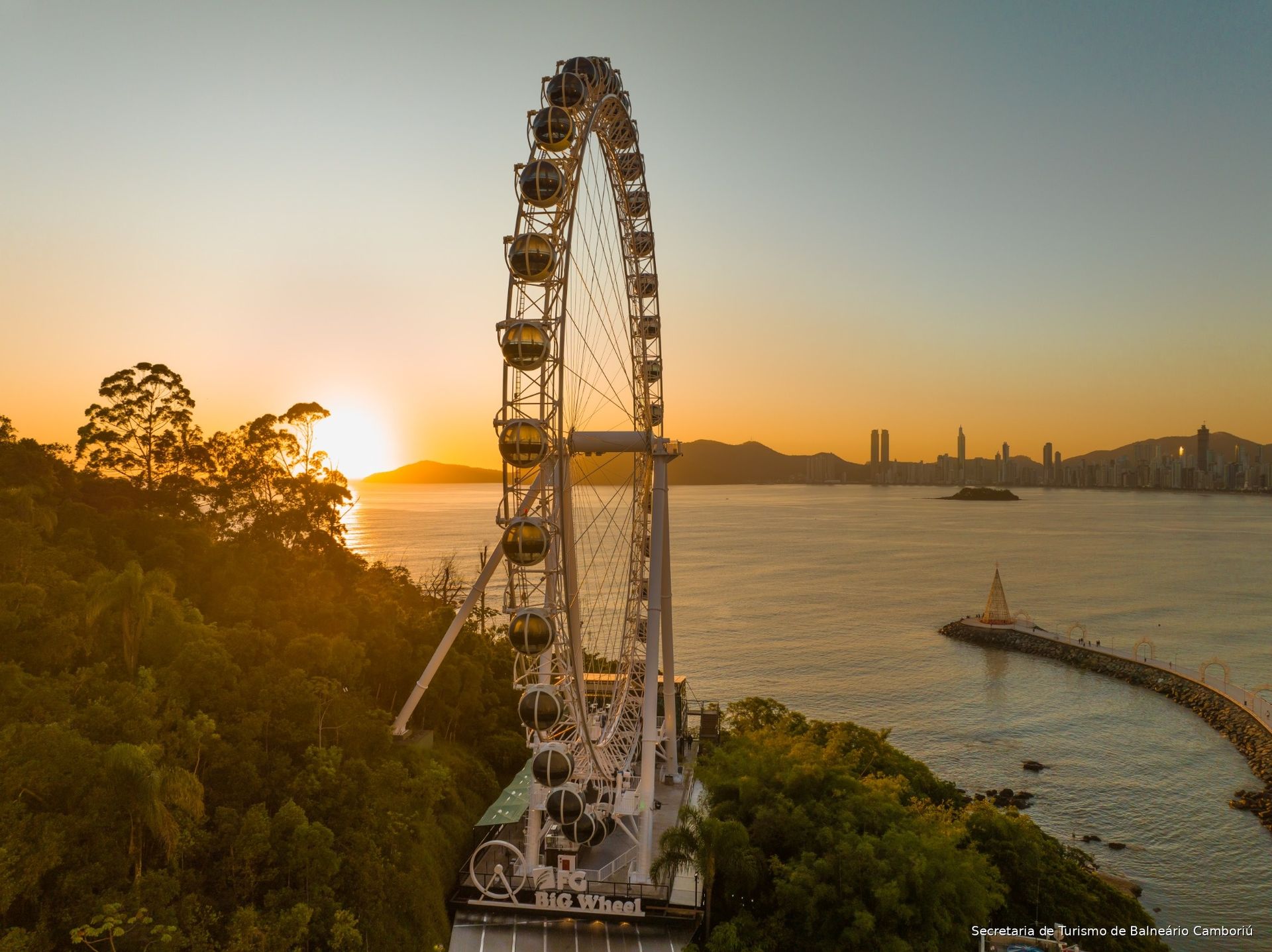 Roda-gigante ao pôr do sol à beira de um rio, com o horizonte da cidade e um pequeno barco na água