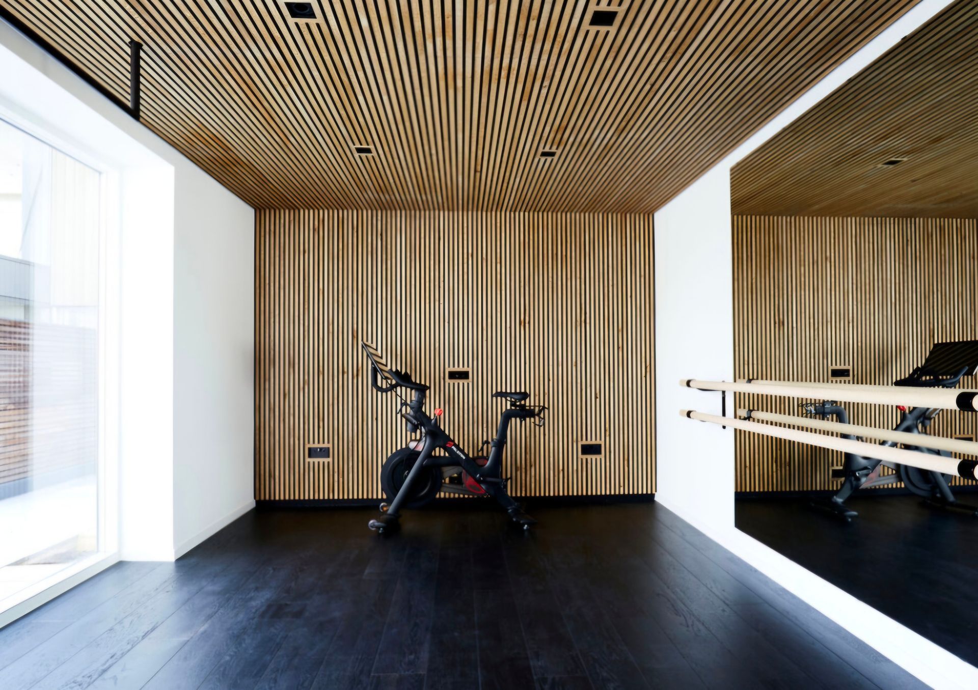 Gym with wooden walls and ceiling, stationary bike, large mirror. Dark wood floor, bright window.