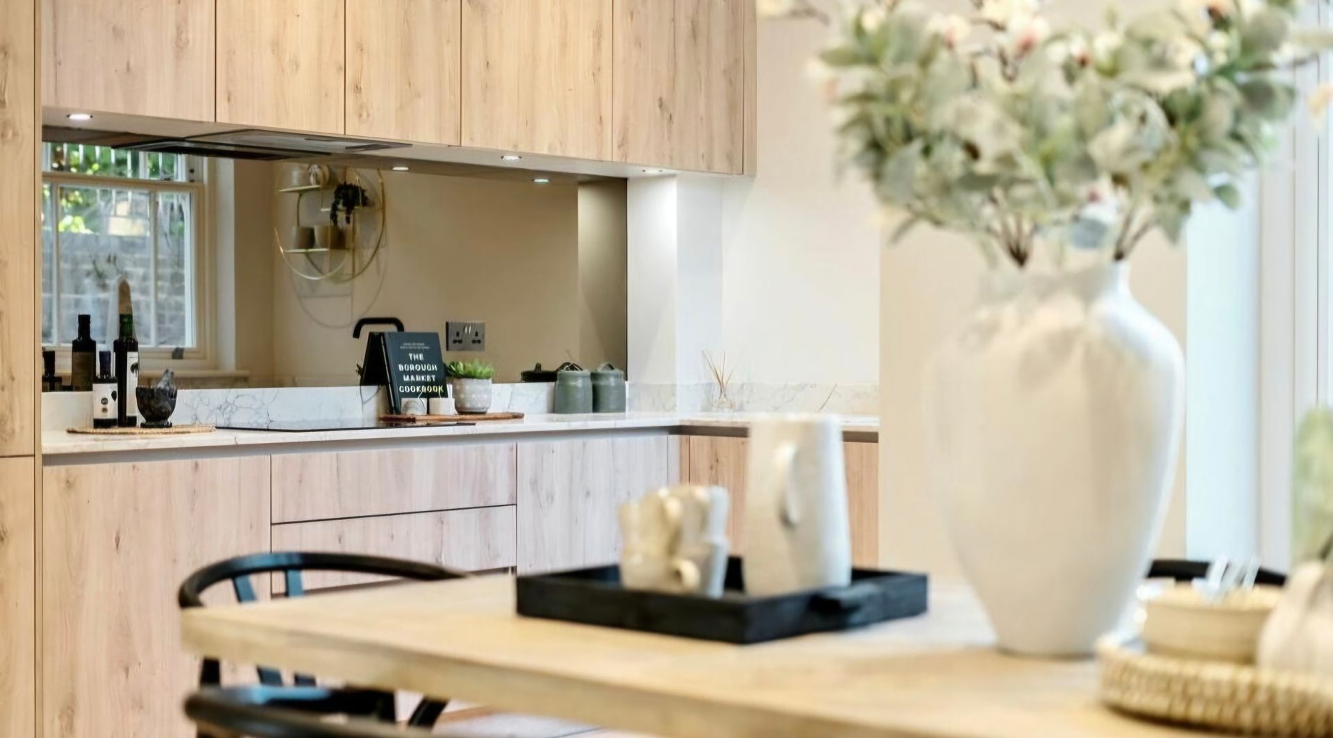 Light wood kitchen with vase of flowers on a dining table in the foreground.