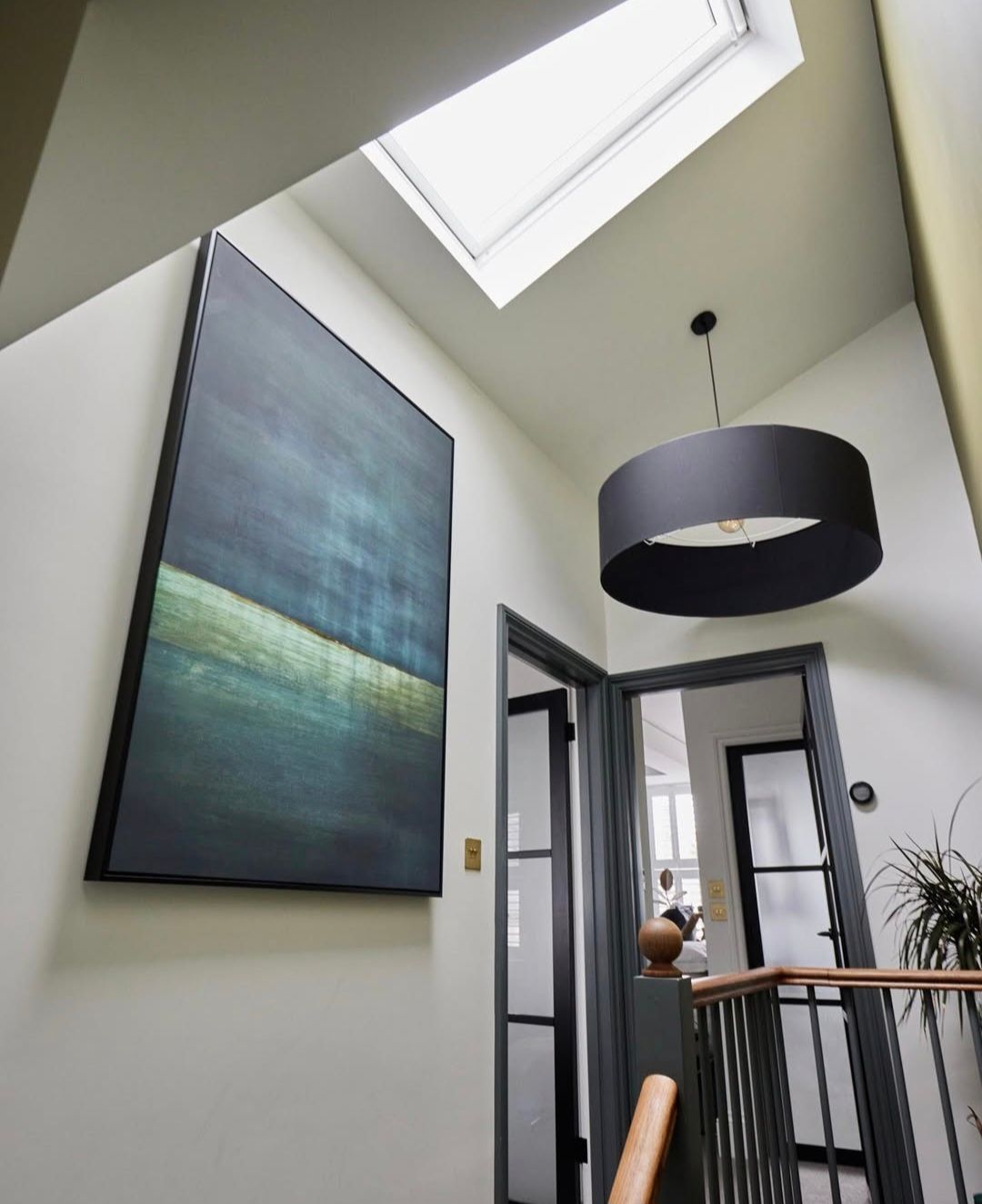 Hallway with large blue-green art, black pendant light, skylight. Dark gray door frames. Staircase with wooden handrail.