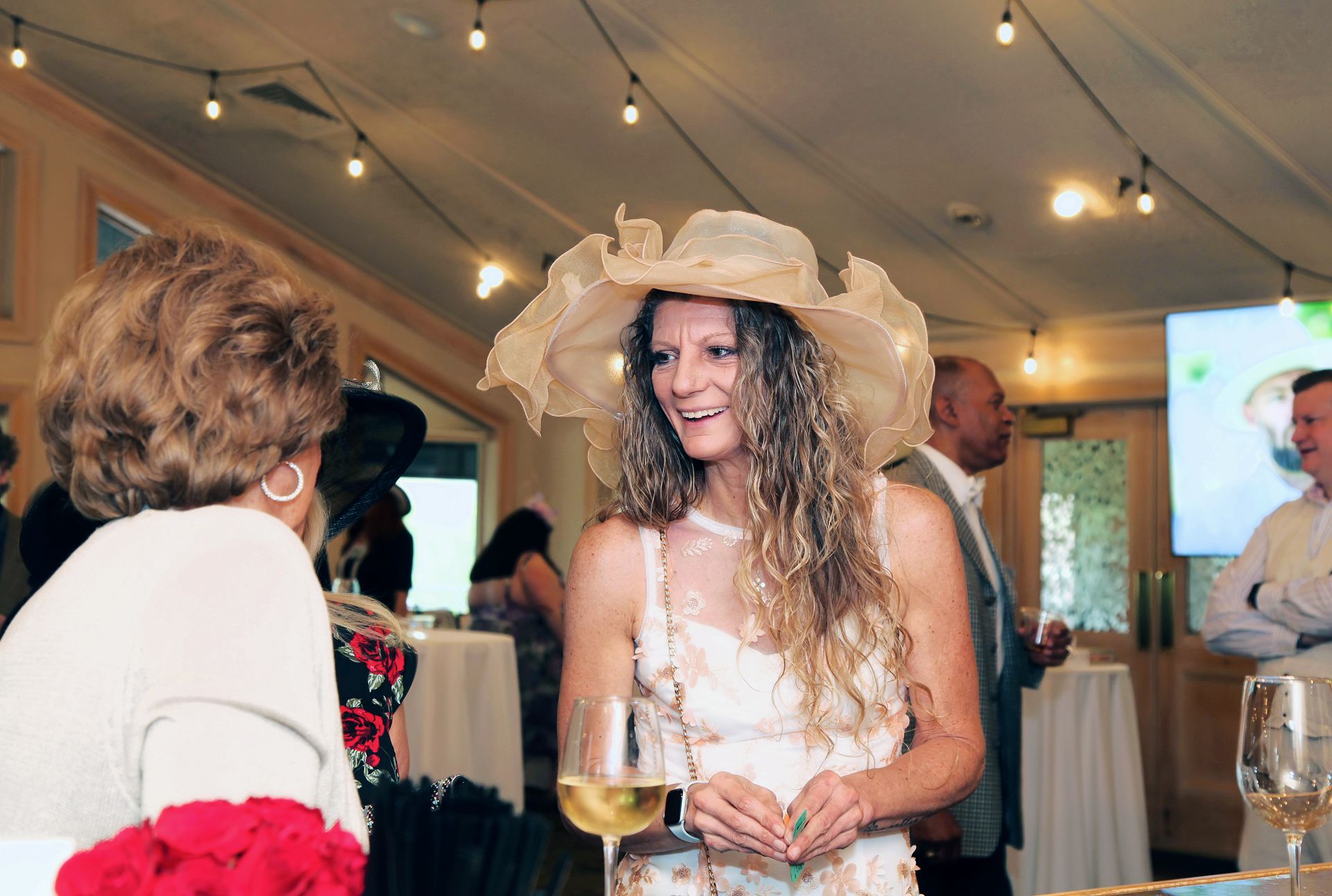 A woman wearing a hat and a white dress is holding a glass of wine.