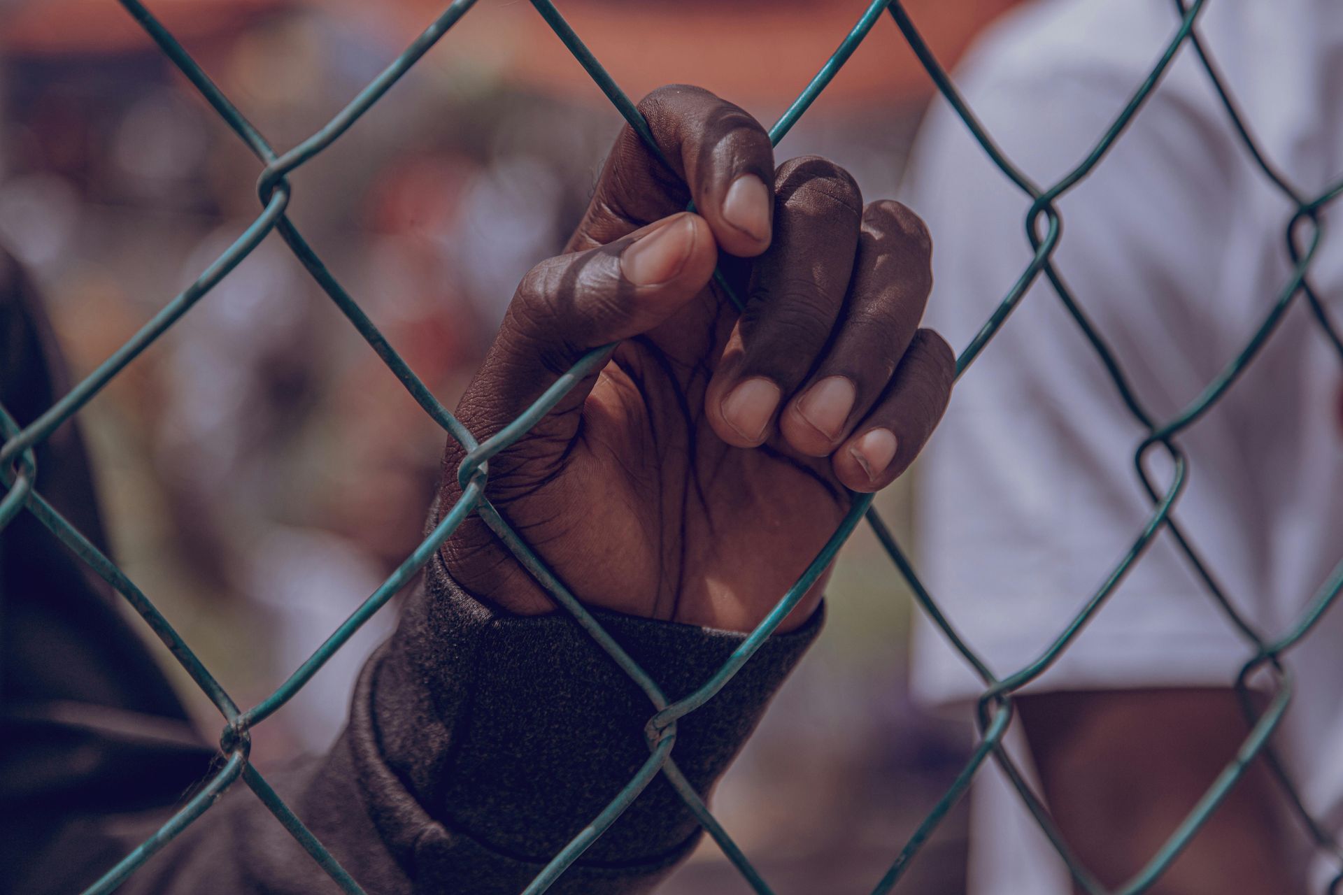 Hands gripping a chain-link fence, with a blurred crowd in the background.