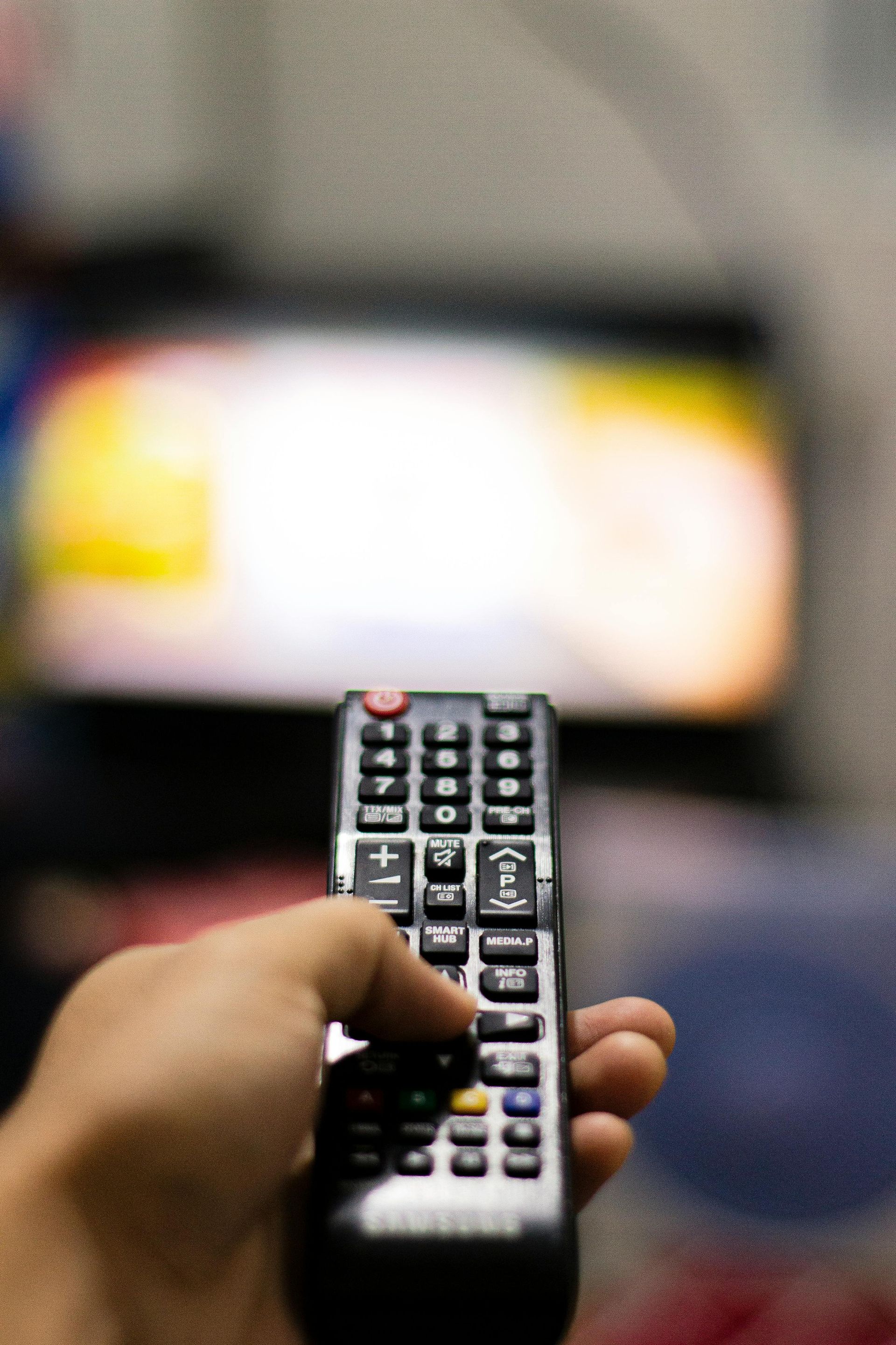 A hand holds a black television remote, pointing it toward a blurry, out-of-focus TV screen in the background.