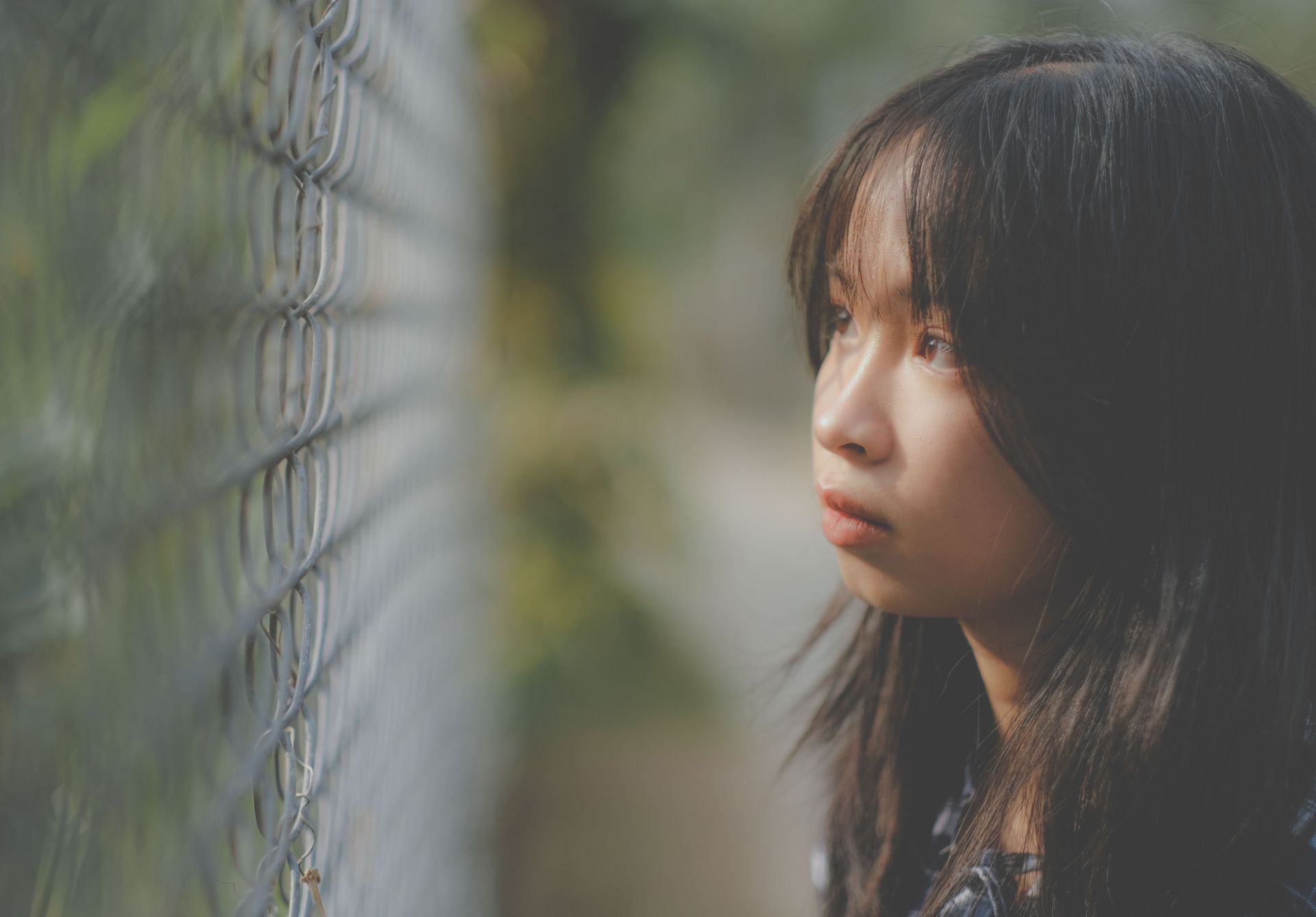 Person in profile gazing through a chain-link fence in soft, blurred outdoor light.