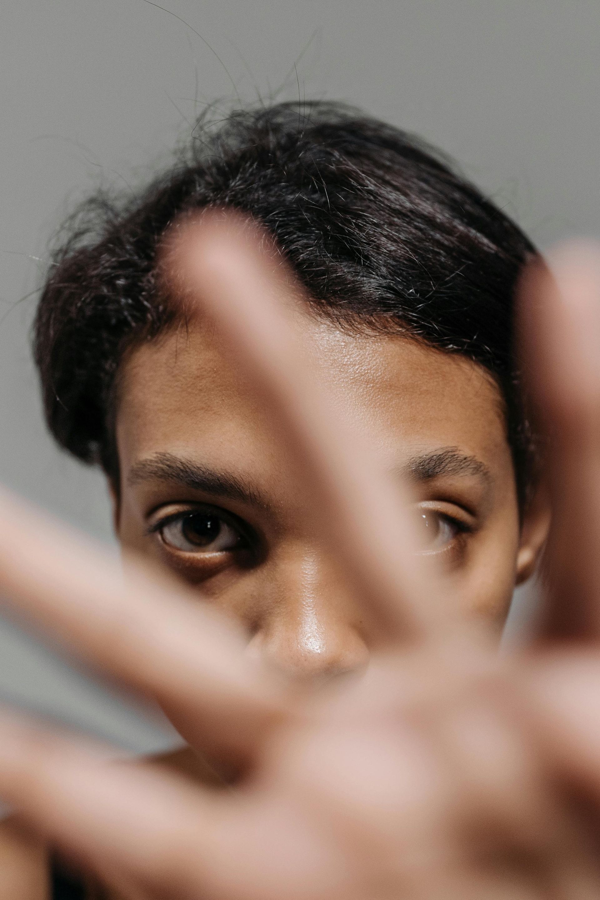Person peeking through raised fingers, face partially obscured against a plain gray background