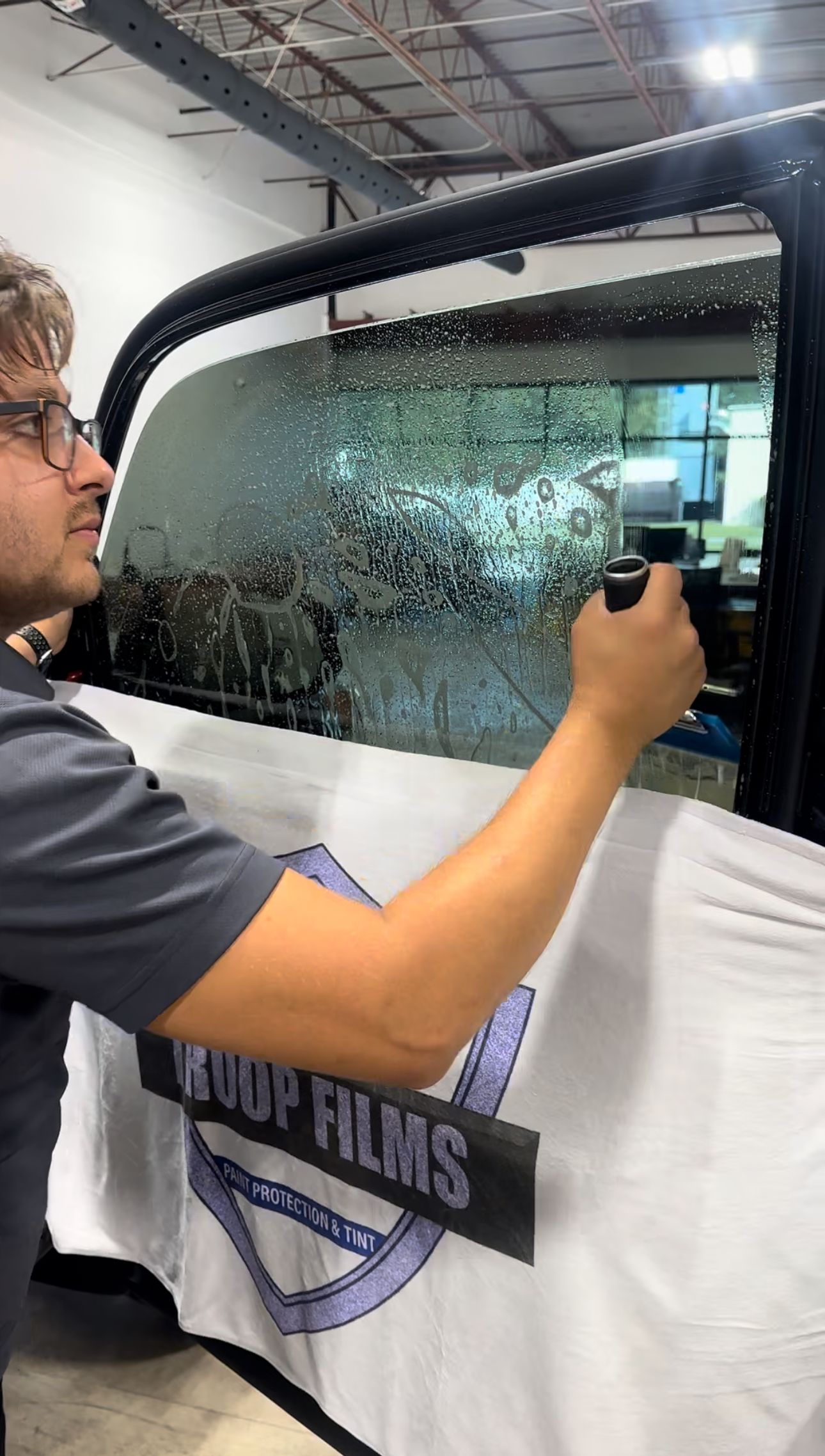 Person applying window tint to a car window; shop setting, using a squeegee.