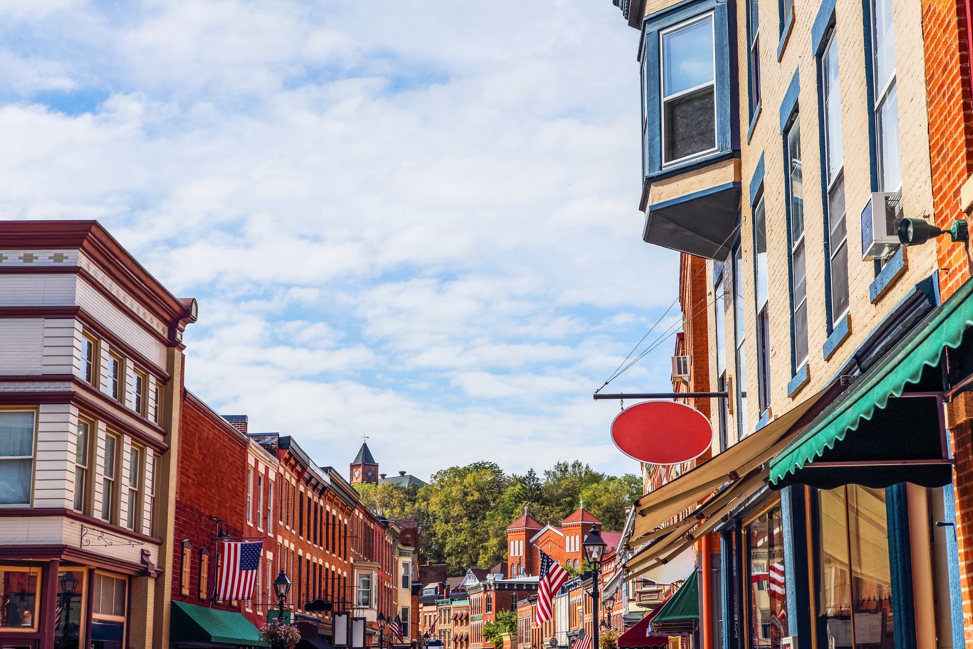 Street lined with brick buildings, shops, and American flags; church on hill in the distance under a partly cloudy sky.