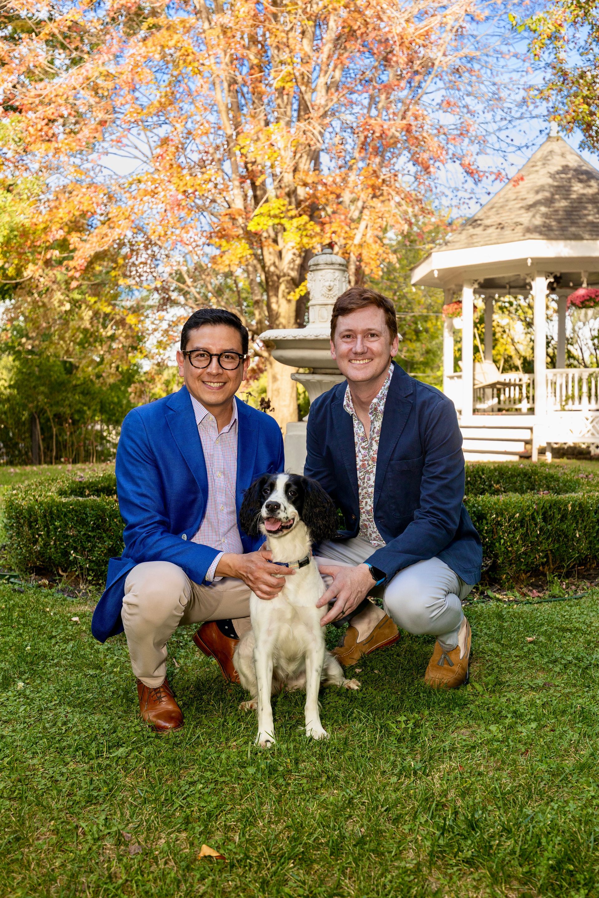 Two men and a dog crouch on grass; autumn trees and a gazebo in the background.
