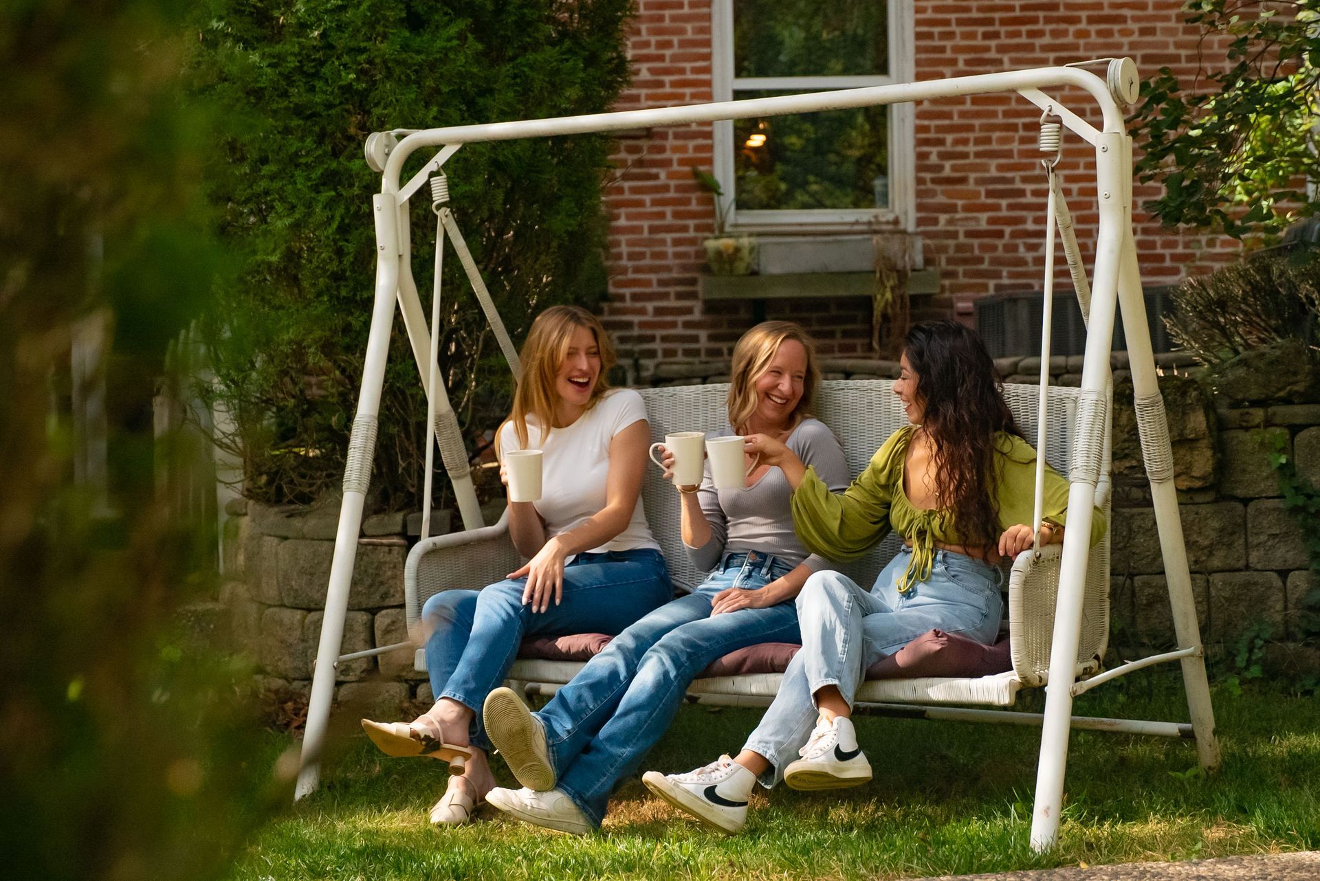 Three people laugh while sitting on a porch swing, holding mugs in front of a brick building.