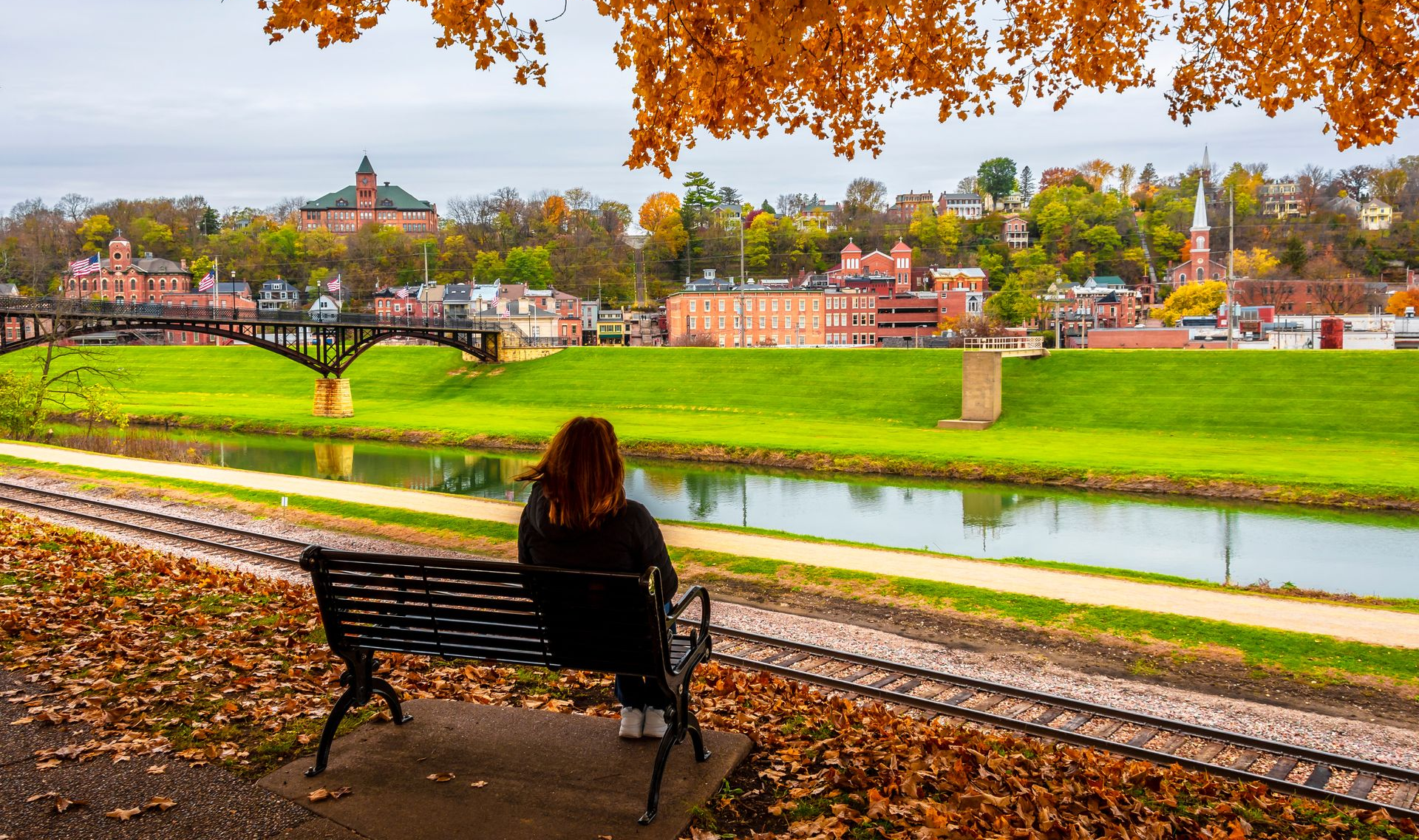 Person sitting on a bench by a river, with autumn trees and colorful houses across the water.