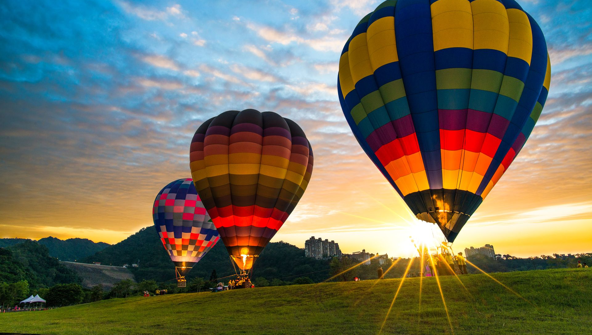 Three colorful hot air balloons floating over a sunrise landscape with mountains and grassland